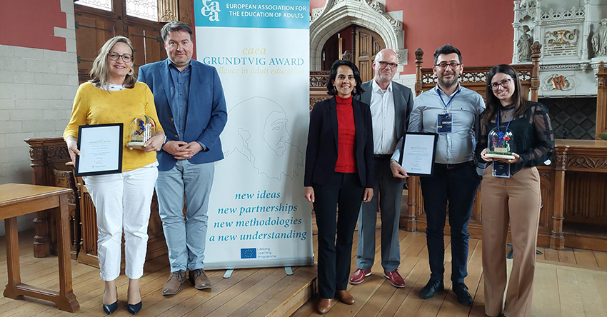 Unterschiedliche Gewinnerinnen und Gewinner des 19. EAEA Grundtvig Awards mit ihren Auszeichnungen stehen vor einem Plakat der Veranstaltung innerhalb eines mittelalterlichen Rathauses in Mechelen, Belgien.